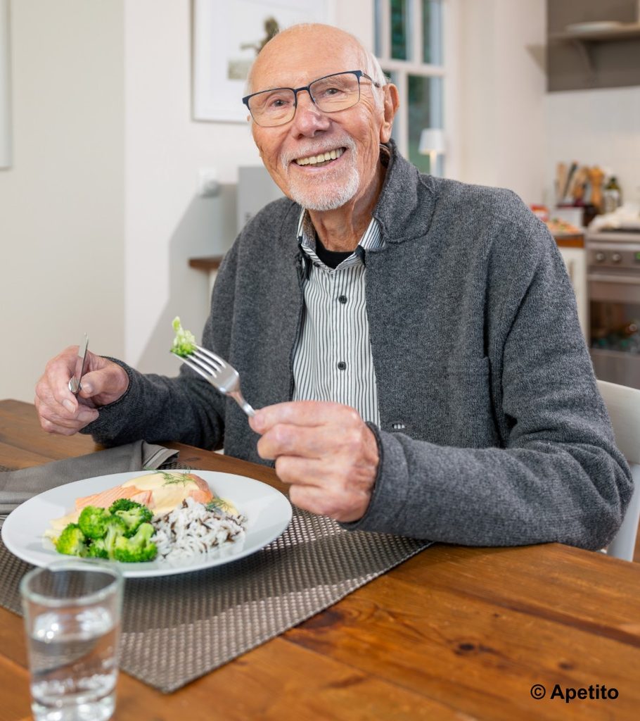 Älterer Mann mit Brille und grauem Pullover sitzt an einem Holztisch und hält Besteck über einem Teller mit Reis, Brokkoli und Lachs